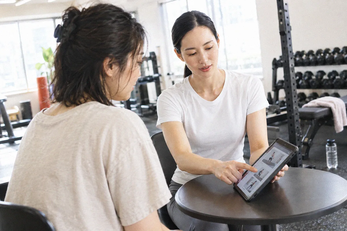 丸テーブルでタブレットの種目画像を指しながら説明するトレーナー
